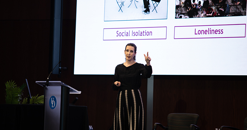 Lisa Jaremka, associate professor of psychological and brain sciences at UD’s College of Arts and Sciences (CAS), holds up two fingers while giving a keynote address on understanding loneliness. Behind her is a projection screen with the words "social isolation" and "loneliness" on it. 