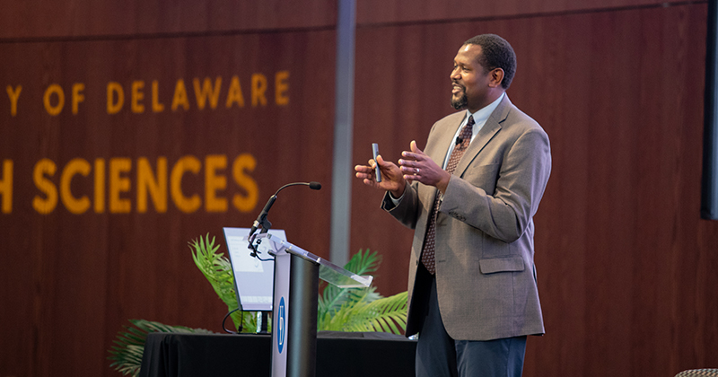 Geriatrician Dr. Thomas Cudjoe, assistant professor of medicine at Johns Hopkins University, gives a keynote address on the epidemic of loneliness. The UD College of Health Sciences is illuminated on the Audion wall around him. 