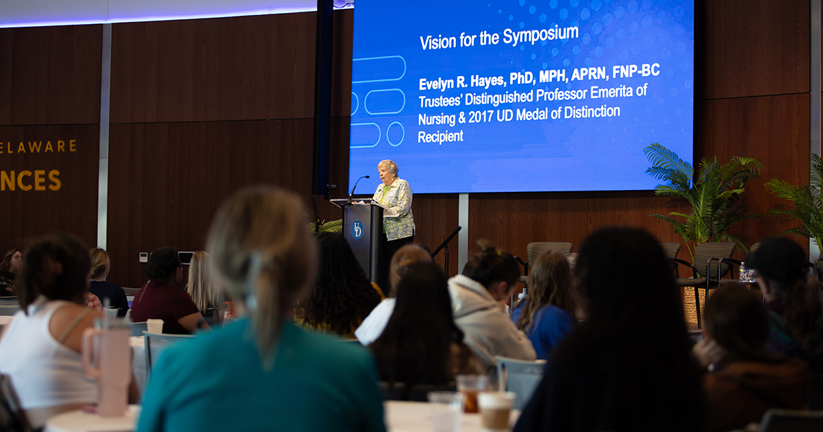Evelyn R. Hayes, Trustees Distinguished Professor Emerita in UD’s School of Nursing, whose generous support makes the annual Hayes Symposium possible, delivers her vision for this year's symposium in a room full of people seated at round tables in the Audion on STAR Campus. Behind Evelyn is a large projection screen with a blue background that reads "Vision for the Symposium" followed by Evelyn's name and title.