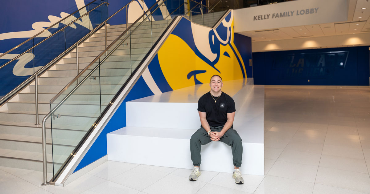 Austin Mark sits on a ledge in the lobby of the Whitney Athletic Center in front of a wall with a giant Blue Hen painted on it