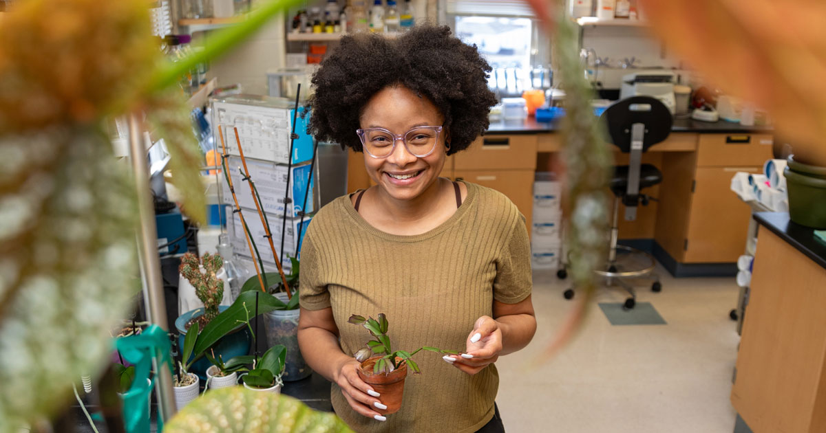 Jazzlyn Jones stands in a Medical and Molecular sciences lab holding a Christmas cactus in a clay pot and surrounded by other plants