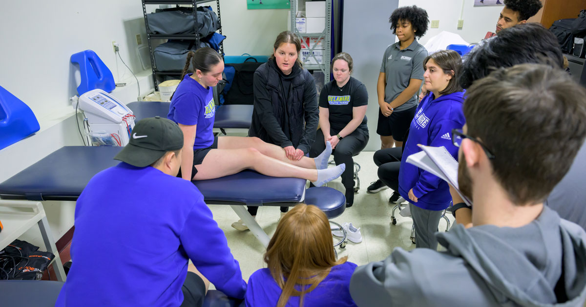 Students stand in a semi-circle around a student on a table with her legs out for a therapeutic modalities class where they're learning about cupping and therapeutic massage.