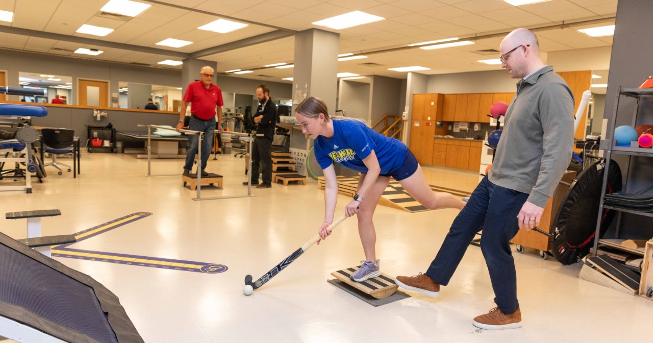 A person holds a field hockey stick and prepares to hit a ball while standing on one leg while another person stands with his foot out as part of a Physical Therapy Clinic exercise.