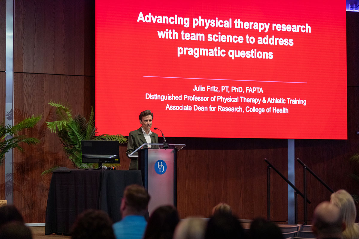 Julie Fritz, distinguished professor of physical therapy and athletic training and associate dean for research at the University of Utah, stands on the Audion stage at a podium with a large orange screen behind her with her title talk's on it, ""Advancing Physical Therapy Research with Team Science to Address Pragmatic Questions."