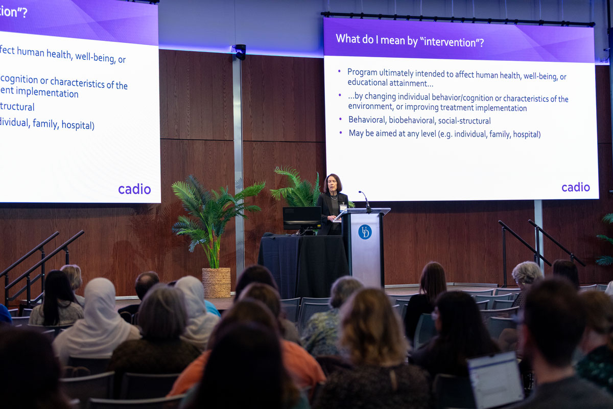 Linda Collins, professor of social and behavioral sciences at New York University, stands at a podium on the Audion stage with a large screen showing her presentation behind her. The screen's topic is "What do I mean by intervention?" and is followed by several examples.