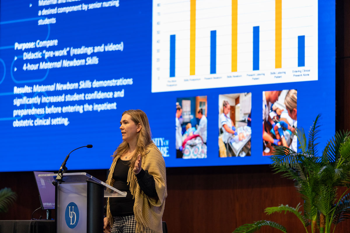 Amanda Watson, assistant professor in the School of Nursing, stans on the Audion stage at a podium to presentp her research on a new skills component for nursing seniors focused on maternal and newborn care. Here, she stands with a blue screen with graphs on it behind her as part of her presentation.