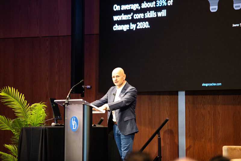 Globally recognized AI expert Alex Goryachev stands at the podium in the Audion at STAR for his keynote speech. Here, he's delivering his presentation on a large black screen with white lettering behind him. 