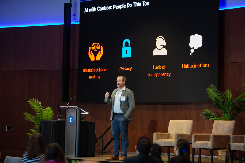 Stern Summit keynote speaker Dan Weberg, executive director of workforce development and innovation at Kaiser Permanente, stands away from the podium delivering his presentation on "Using AI to Optimize Clinical Workflow." His presentation on a large black screen with colorful icons is behind him.  With each icon are the words, "Biased decisionmaking," "Privacy," "Lack of Transparency," and " Hallucinations." 