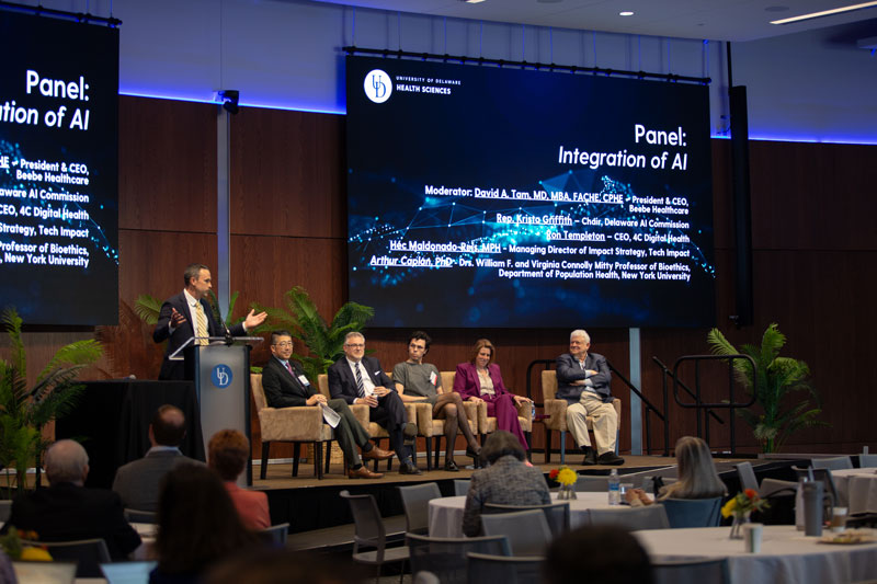 Mike Smith, (left), director of strategic initiatives for CHS, introduces a panel on the "Integration of AI" moderated by David Tam, president and CEO of Beebe Healthcare, and panelists (L to R): Ron Templeton, CEO of 4C Digital Health; Héc Maldonado-Reis, managing director of impact strategy for Tech Impact; State Rep. Krista Griffith, chair of the Delaware AI Commission; and Arthur Caplan, Drs. William F. and Virginia Connolly Mitty, Professor of Bioethics in the Department of Population Health at NYU.  Behind them is a blue screen that reads: "Panel: Integration of AI" and features the moderator and panelists' names in white italics.