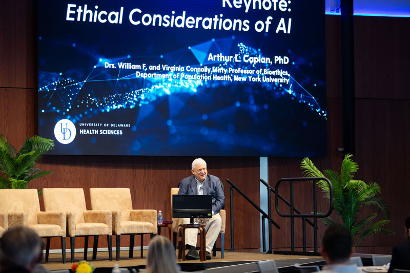 Stern Summit keynote speaker Arthur Caplan, Drs. William F. and Virginia Connolly Mitty, Professor of Bioethics in the Department of Population Health at New York University,  is seen seated giving his presentation on the stage in the Audion at STAR. Behind him is a large blue screen that reads: "Keynote, Ethical Considerations of AI" and includes his name and title. 