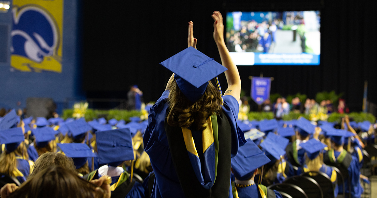 A sea of blue caps taken from behind with one student standing to applaud at the CHS Convocation. A Blue Hen flag is in the corner, and a TV screen showing Convocation is in the front atop the stage.