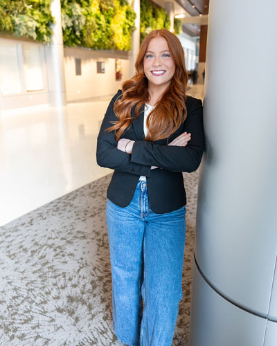 Senior health behavior science major Emily Gallo leans on a pole with her arms crossed wearing a black blazer and jeans for a posed photo with the Living Wall on STAR Campus behind her.