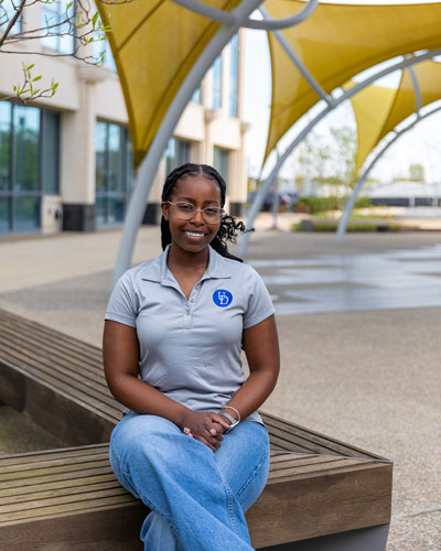 Zenas Okera, a senior honors nutrition and medical sciences major, sits on a bench outside on STAR Campus. She's wearing a grey shortsleeve UD shirt and blue jeans.