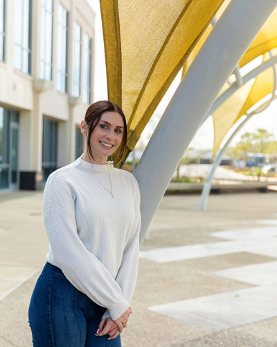 Senior applied molecular biology and biotechnology major Victoria Barbone poses outside STAR Campus wearing a white sweater and blue denim pants.