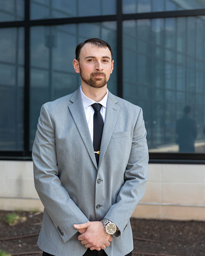 Senior Shane McGarry, a medical laboratory science major, wears a light grey suit and stands with his hands clasped as he poses for a headshot outside on STAR Campus.