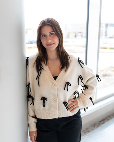 Senior medical laboratory science major Bettina Bauman stands against a pole with a hand on her hip wearing a white sweater with black draping bows and black pants with the windows of the STAR Atrium behind her..