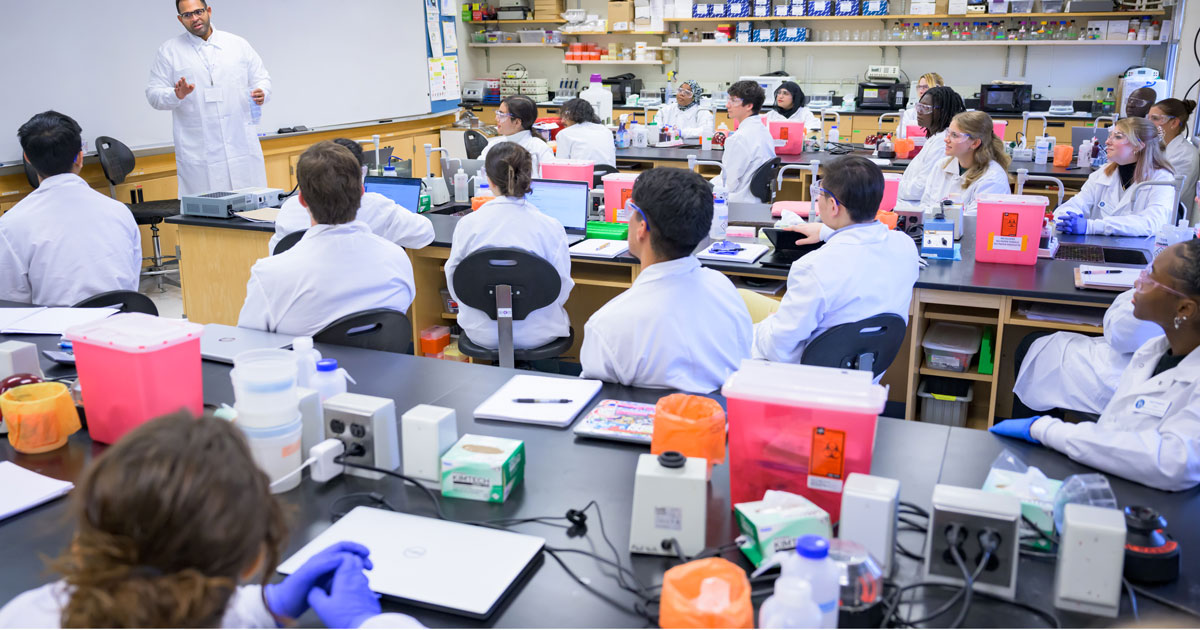 A representative o QIAGEN gives a lecture in a lab to MMSC students who are wearing white lab coats and have instruments ready to use on lab tables.