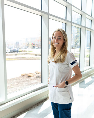 Headshot of Ashley Hommel wearing her white UD nursing shirt and blue scrubs and her nametag pinned at the bottom. The glass windows of the STAR Campus lobby are behind her.