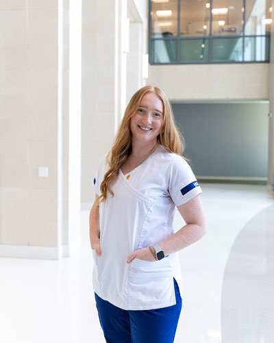 Senior nursing major Emma Oconowicz wears her white nursing shirt and blue scrubs and poses for a headshot with her hands in her pockets in the STAR lobby.