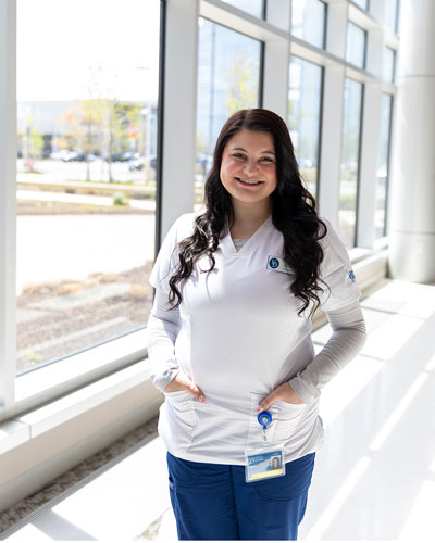 Headshot of senior nursing major Samantha Muza wearing her white UD nursing shirt and blue scrubs and her nametag pinned at the bottom. The glass windows of the STAR Campus lobby are behind her.