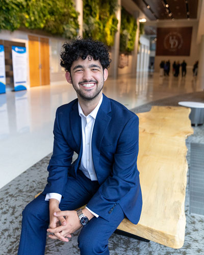 Jackson Tomasco wears a blue suit and a white shirt and sits with his hands clasped on a bench with the Living Wall behind him in the STAR Atrium.