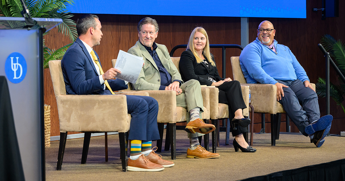 (L to R seated in beige chairs) Michael Smith, director of strategic initiatives for the College of Health Sciences, moderates a panel on resiliency featuring Chris Locke, founder of SL24 and Sean’s House; Cynthia Diefenbeck, director of UD’s Center for Counseling and Student Development (CCSD); and Gregory Cooper, associate director of psychiatric and addiction services at the CCSD, at the 2025 Evelyn R. Hayes Innovations in Healthcare Symposium.  A blue UD background is behind them, and a UD-branded podium is next to Mike Smith.