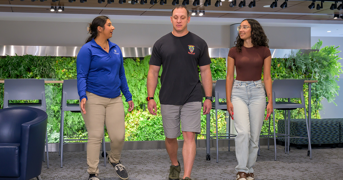 Graduate students work with a participant to promote healthier lifestyle habits. Here they are showing mid-walk with the greenery of the Living Wall behind them.