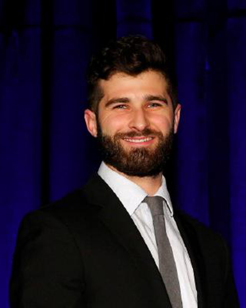 A headshot of Jonathan Wood, assistant professor of physical therapy, wearing a suit and tie with a dark blue background.