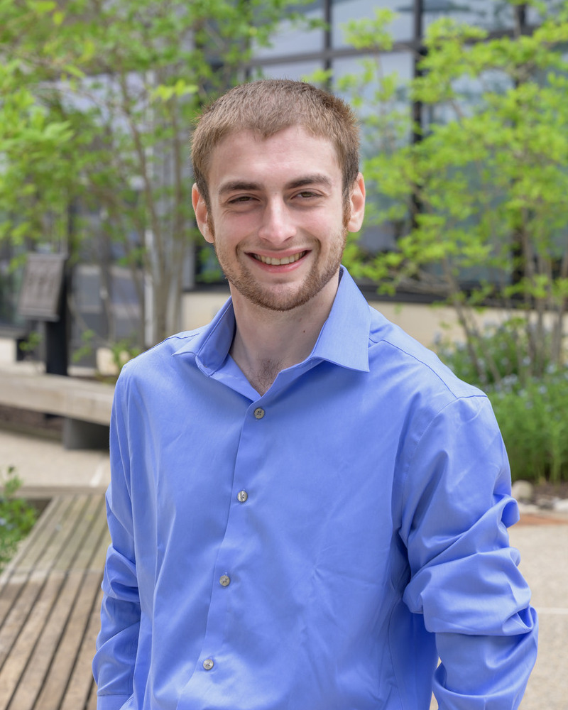 A headshot of Eric Dombrower, a senior medical laboratory science major, wearing a blue buttondown shirt. He's outside with greenery behind him. 