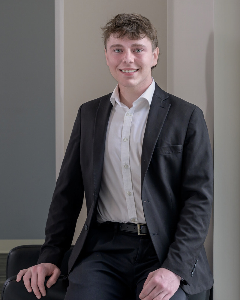 A headshot of John Arnold, a senior medical diagnostics major, is seated wearing a black jacket nad white shirt with his hands on his knees.