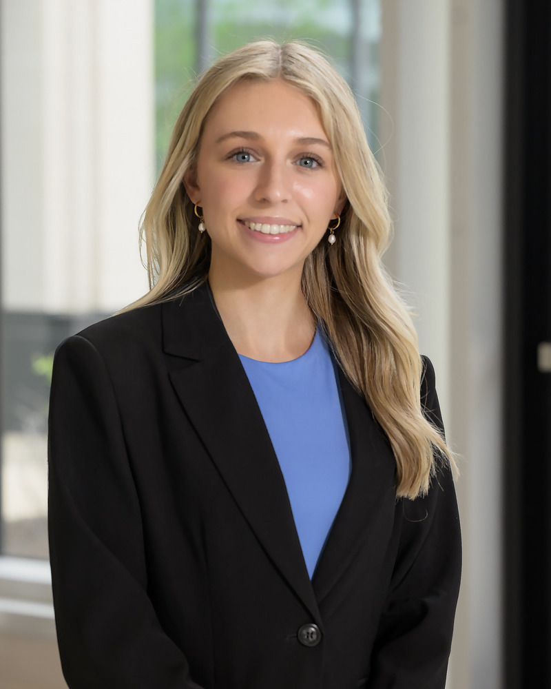 A headshot of Kylee Leverage, a senior medical diagnostics major with a pre-physician assistant concentration, is wearing a black blazer with a blue shirt underneath with windows behind her.