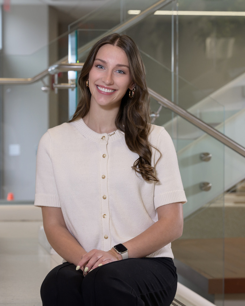 A headshot of Sarah Buckingham, a senior medical diagnostics major with a pre-physician assistant concentration, is seated with glass stairs behind her. She's wearing a buttondown off-white, short-sleeved blouse and black pants. 