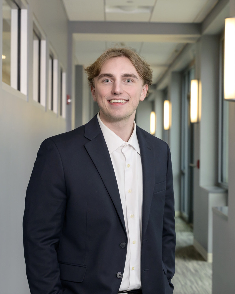 A headshot of Sean Fletcher, a senior medical diagnostis major, wearing a black jacket and a white shirt standing in a long hallway