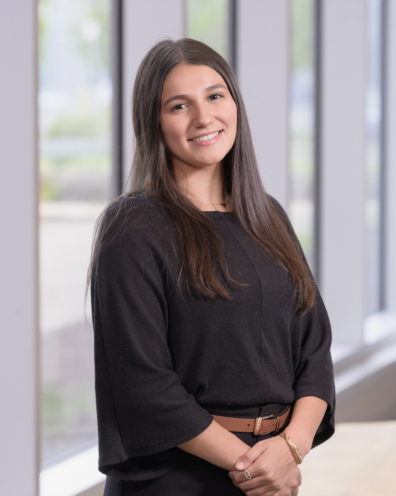 Sophia Lopez,  senior medical laboratory science major, stands with her vack to windows and her hands clasped in front of her. She's wearng a black top with a brown belt.