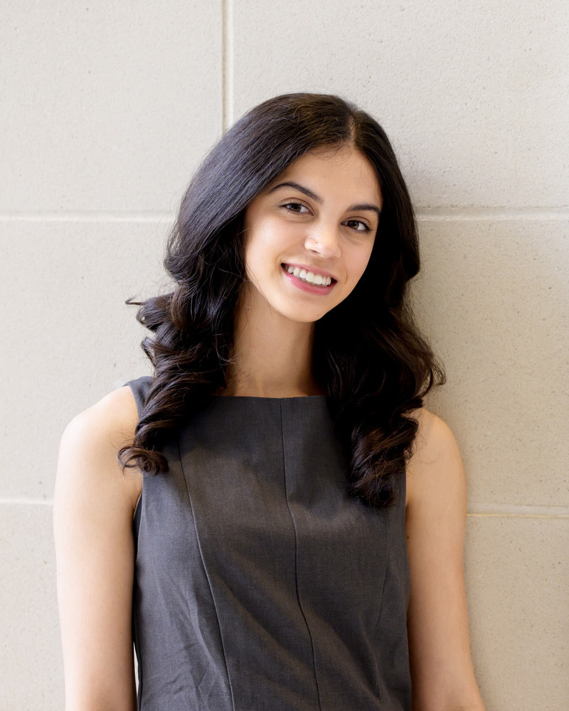 A headshot of Dimple Swain, a senior medical diagnostics major with a pre-physician assistant concentration,  wearing a black sleeveless top with a white tiled wall behind her. 