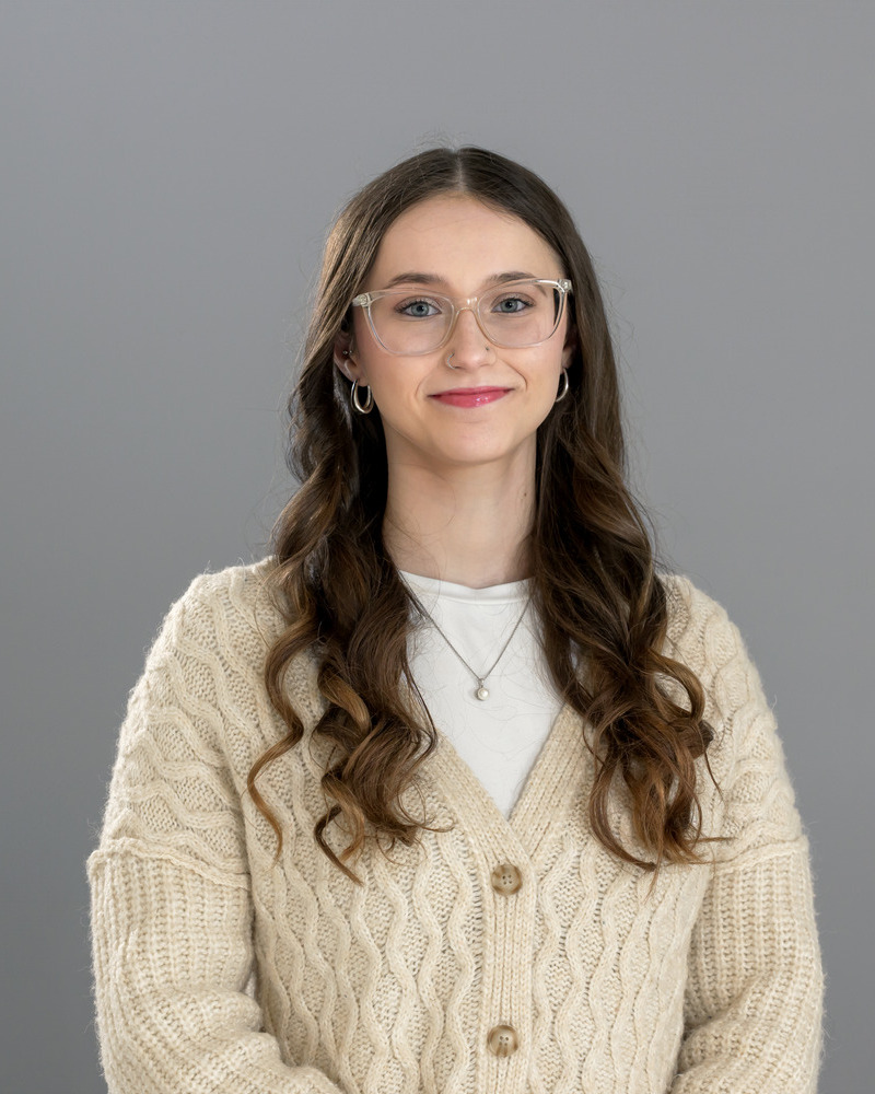 A headshot of Kimberly Salamatin, a medical laboratory science major, is wearing a cream buttondown sweater with a white shirt underneath, with a gray background. She has long curly brown hair and glasses.