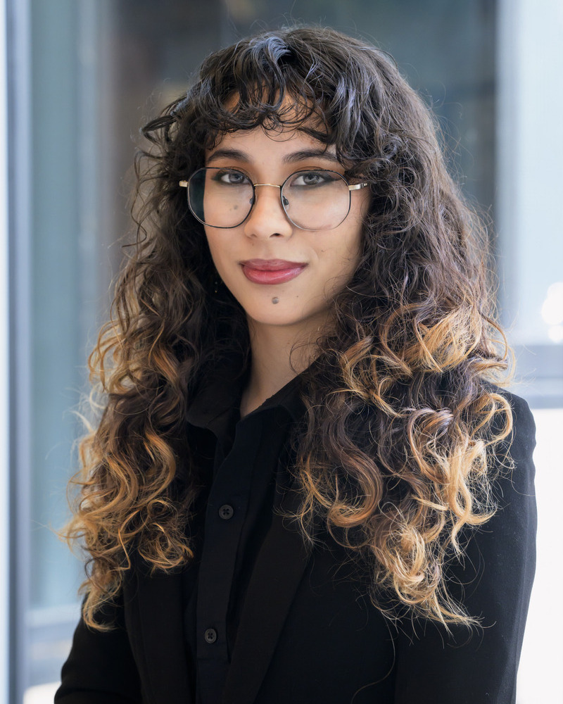 A headshot of Isabel Wilson, an honors medical diagostics major,. She has glasses and curly hair and is wearing a black, long-sleeved top.
