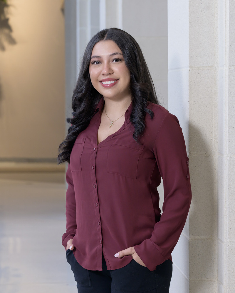 A headshot of Shelsie Martinez, a senior medical diagnostis major with a pre-physician assistant concentration stands with her hands in her pockets. She is wearing a maroon buttondown blouse and black pants. 