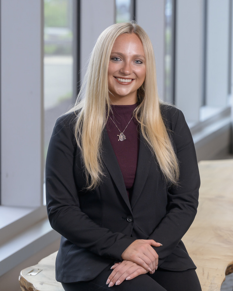 A headshot of Aubrey Menold, a senior medical diagnostics major with a pre-physician assistant concentration, seated with windows behind her. She's wearing a black blazer and a maroon shirt. 