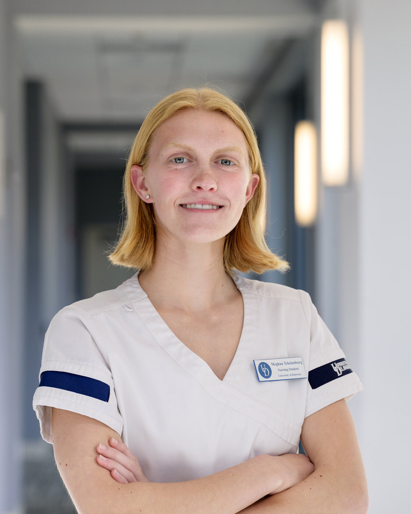 A headshot of Meghan Tekelenburg, a senior nursing major, wearing white scrubs and her nametag with her armed across her chest. She's standing in a well-lit hallway. 