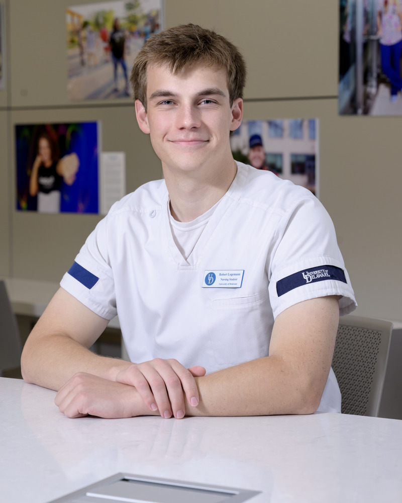 Robert Logemann, a senior nursing major, wears his scrubs and a nametag while seated at a table with one arm over the other. 