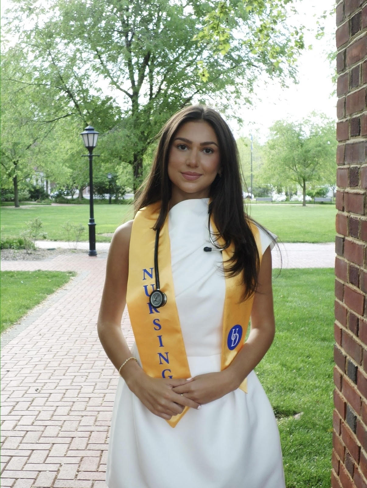 Ava Bonarrigo, a senior nursing major, poses outside on UD's Campus for a senior portrait wearing a white dress and a yellow silk sash that says NURSING in blue capital letters on it. She also has a stethoscope around her neck and her hands clasped.