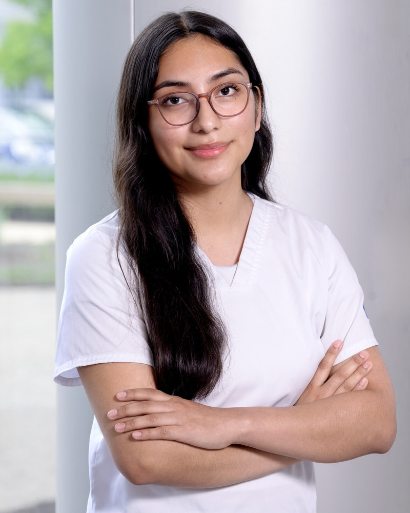 Joanna Cap-Ortiz, a senior nursing major, wears white scrubs and stands outside with her arms crossed over her chest against a large white pillar.