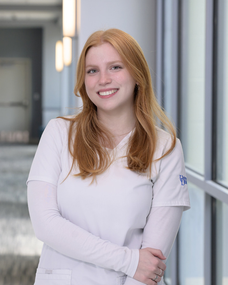 A headshot of Lillian Whitesell, a senior nursing major, standing along a wall of windows with one arm across her chest holding her other arm. She's wearing white scrubs and has strawberry blonde hair just past her shoulders.