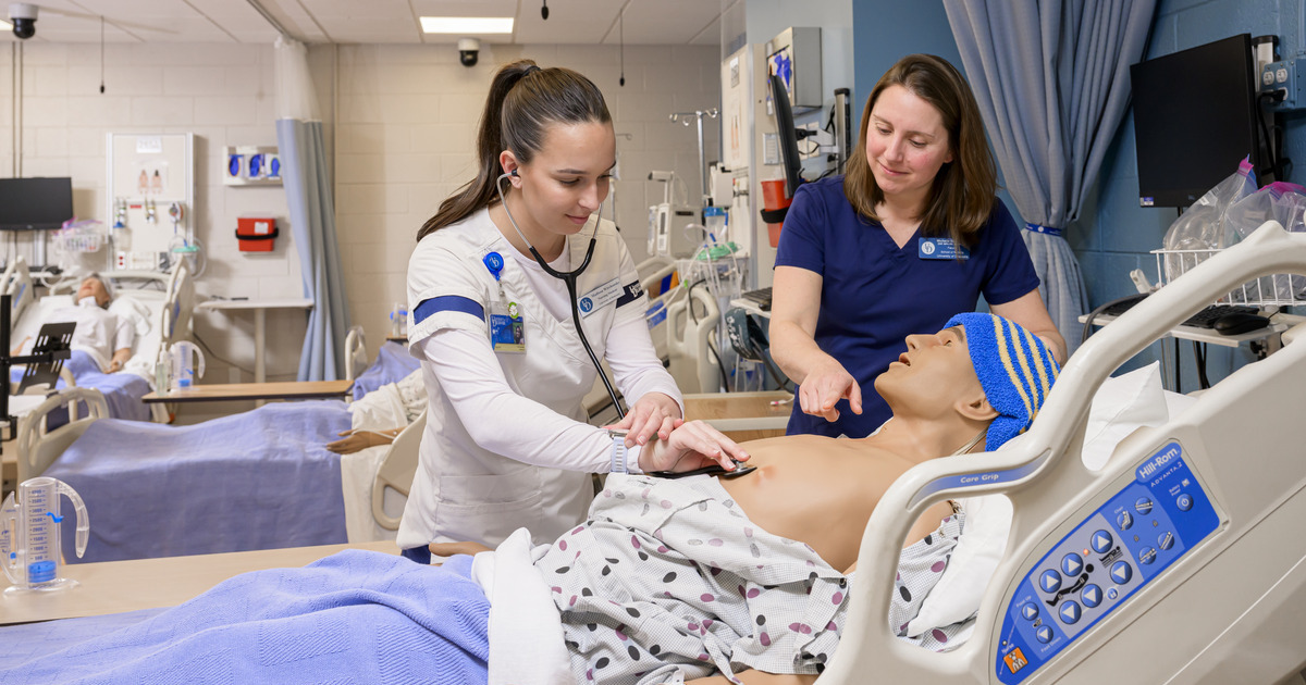 School of Nursing senior Madison Wirchansky uses a stethsoscope and looks at her watch to listen and time out breaths on a high-fidelity manikini in a patient bed while Assistant Professor of Nursing Michelle DePhillips supervises..