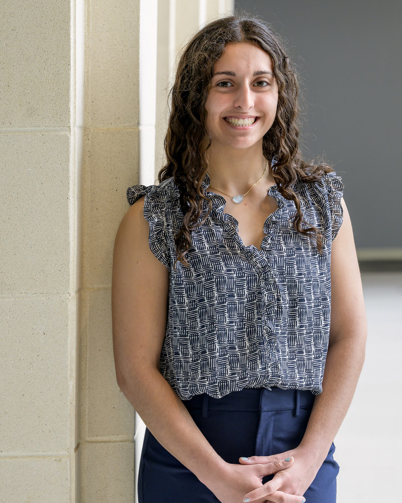 A headshot of senior Adriana Mazzella standing against a wall with her hands clapsed. She's wearing navy trousers and a black and white sleeveless ruffled top. 