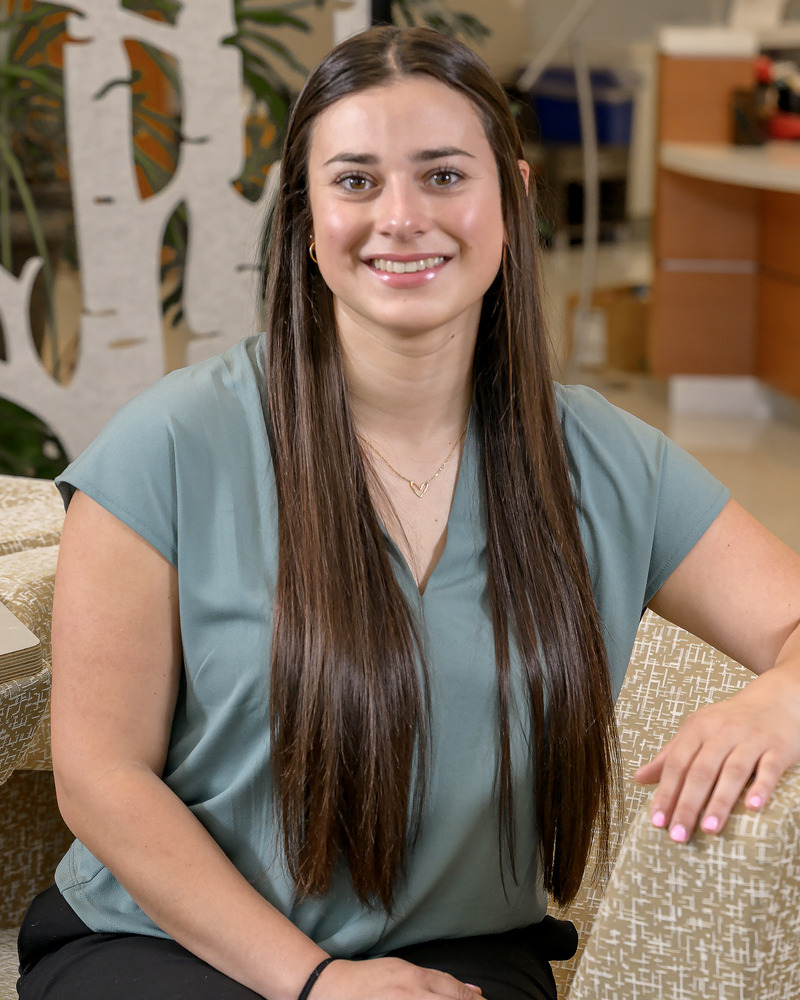 Senior Keuka Miranda Wiltberger is seated with her arm on a chair wearing a sage green v-neck shirt and dark pants. 