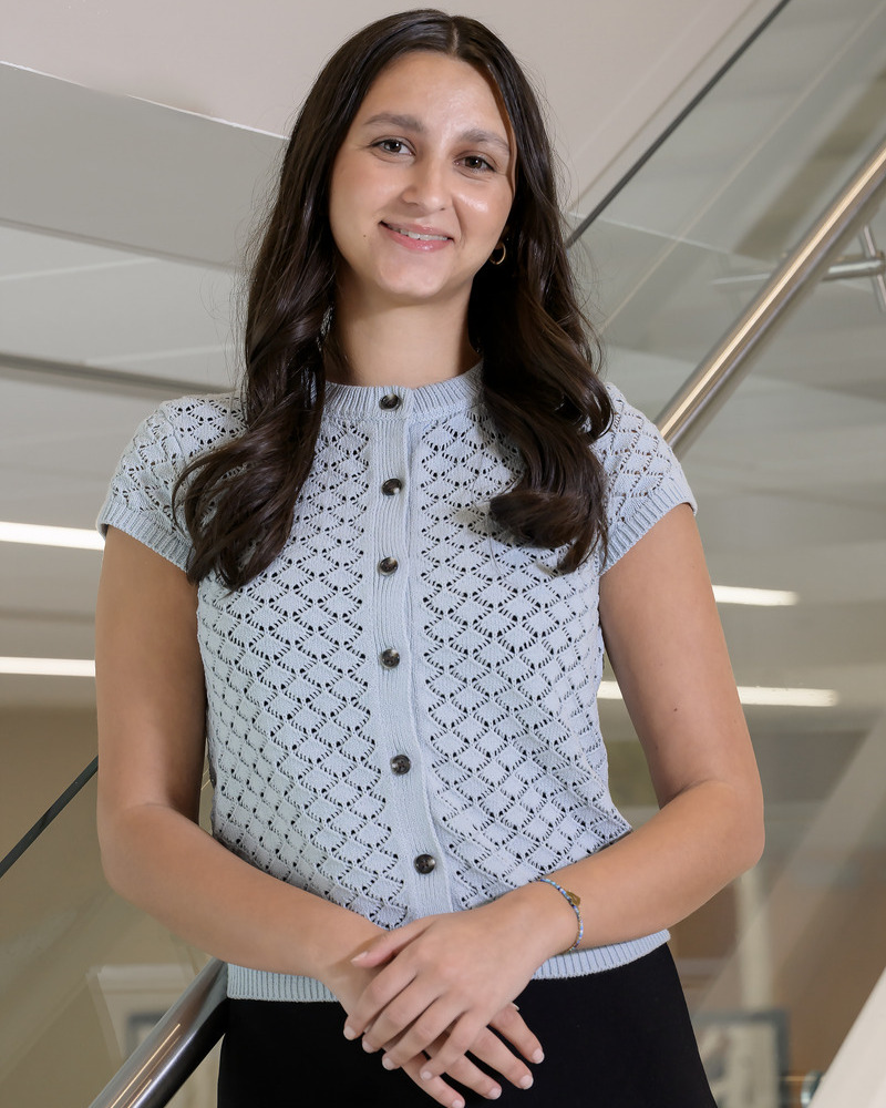 A headshot of senior honors health behavior science major Ellery Bergman Chudnow stading on stairs with her hands clasped. She's wearing a light blue patterened button down sweater