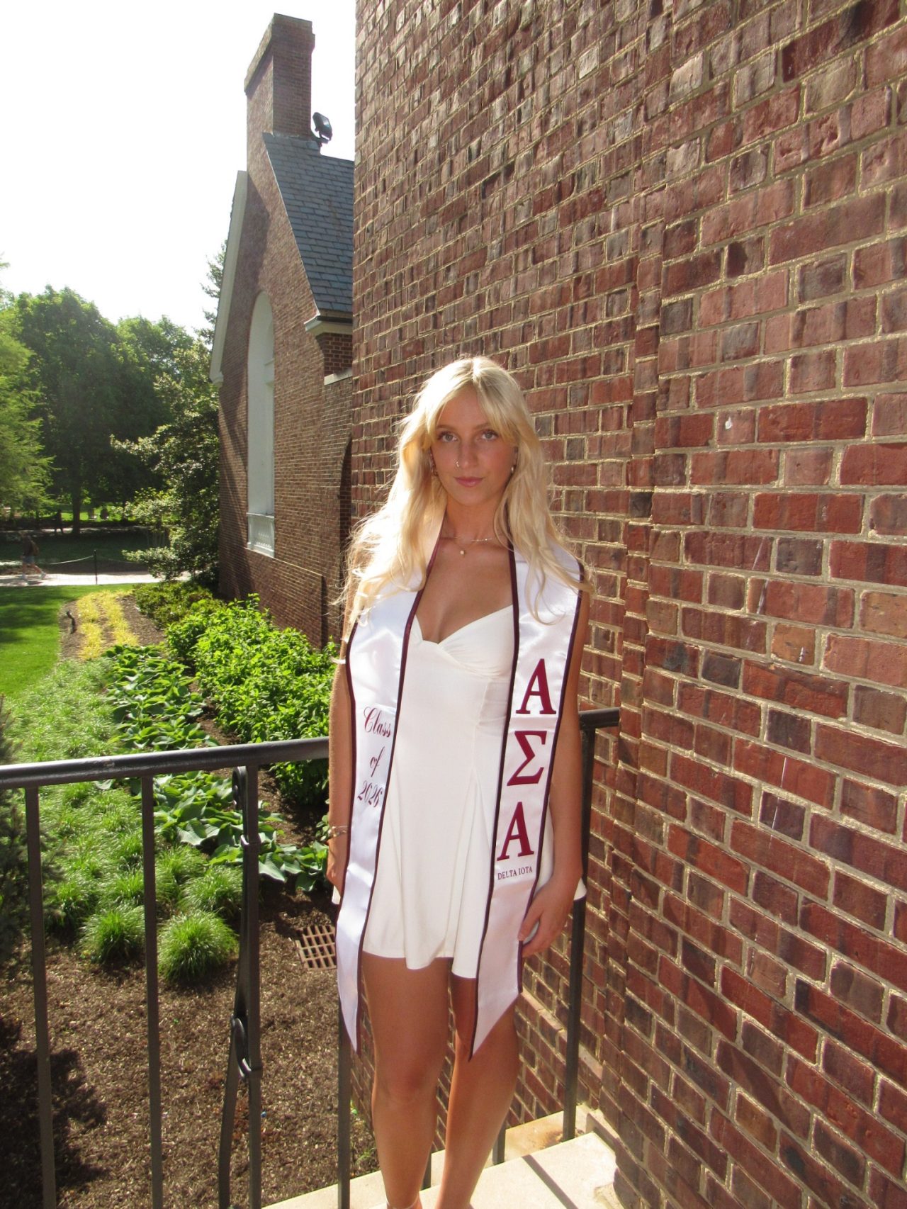 A headshot of senior health behavior science major Taylor Smeby wearing a white dress with her sorority silk sash with a brick building behind her.
