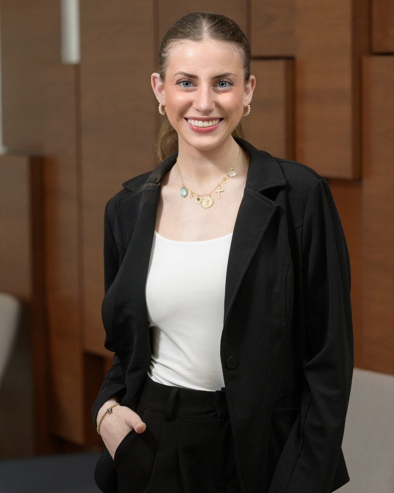 Bella Cipolloni, a senior kinesiology major, poses for a headshot wearing a black suit jacket and a white shirt with her hands in her pockets and her hair pulled back, with a brown wood wall behind her.